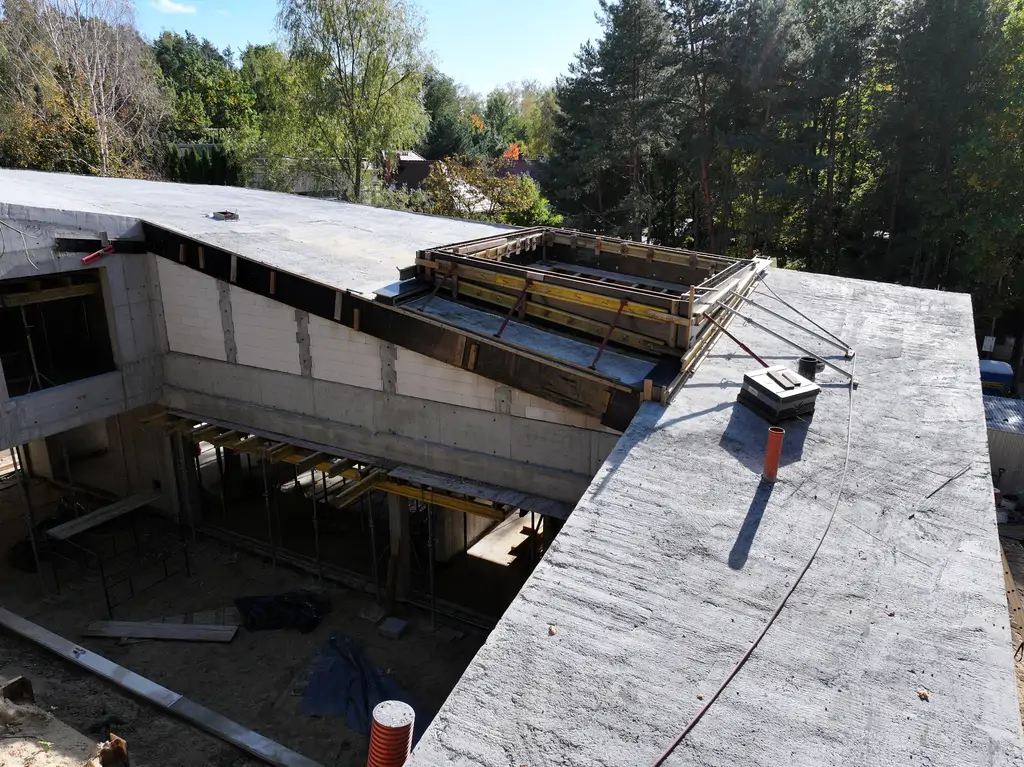 Widok na żelbetowy dziedziniec nowoczesnego domu z pochyłą bryłą, otwartym patio i pracami konstrukcyjnymi w toku.