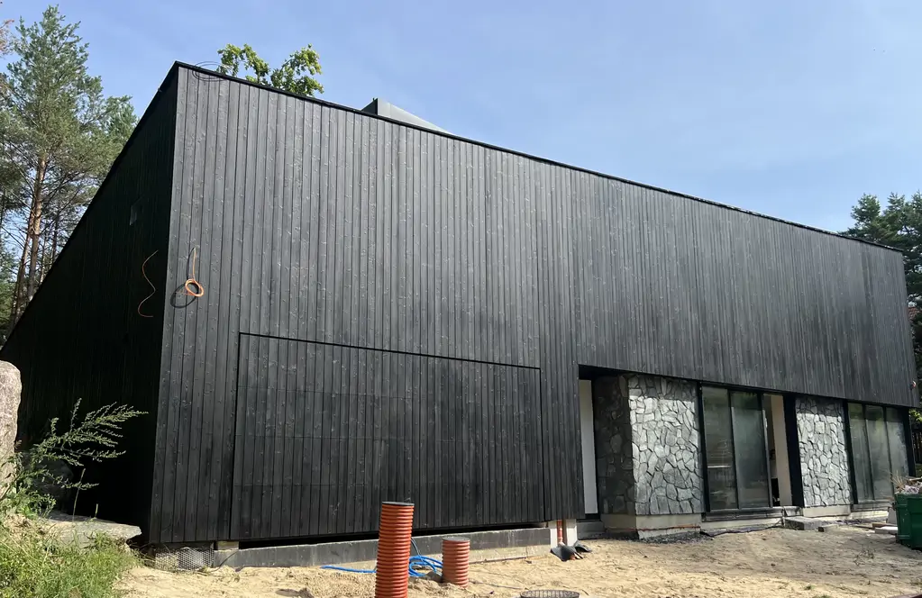 Frontowa elewacja nowoczesnego domu z czarną drewnianą okładziną, kamieniem naturalnym i dużymi przeszkleniami.