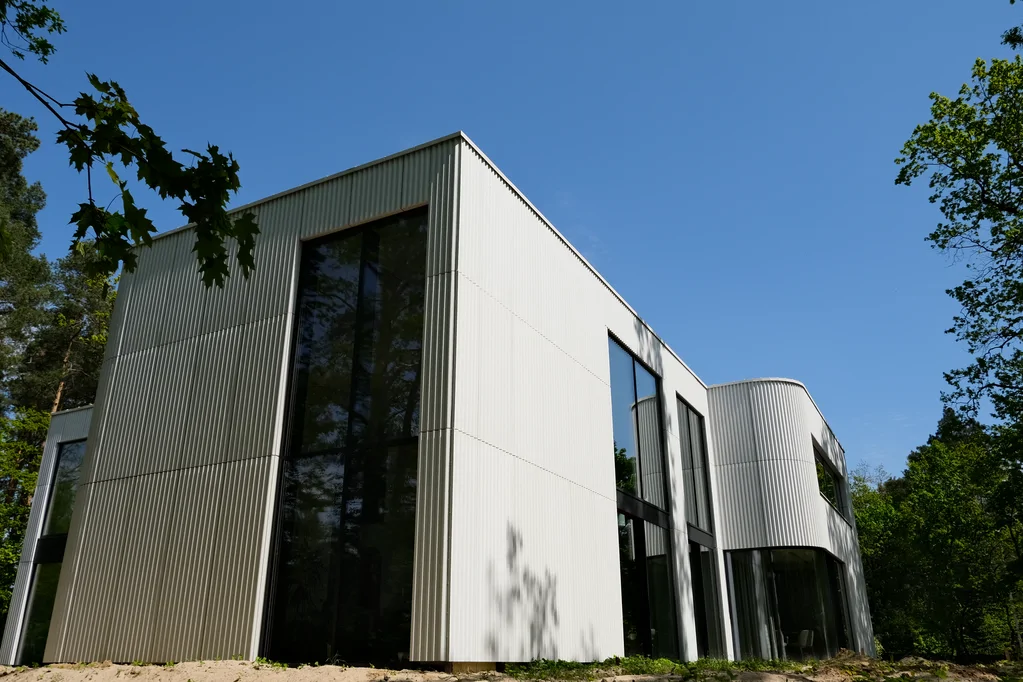 Boczna elewacja nowoczesnego domu z wysokimi przeszkleniami, jasną ryflowaną okładziną i zaokrąglonym narożnikiem w leśnym otoczeniu