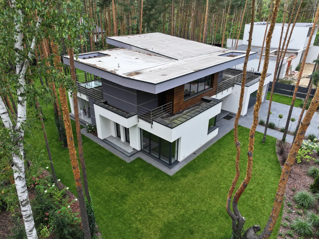 Widok z drona na tył domu: duże okapy, taras i przeszklenia, zielony trawnik między sosnami