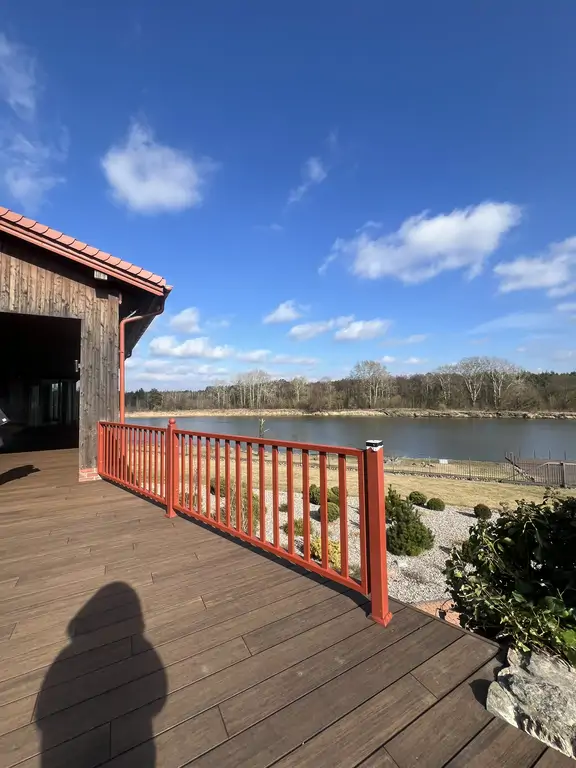 Taras po remoncie z widokiem na rzekę, czerwonymi balustradami i drewnopodobną posadzką przy drewnianej elewacji domu.