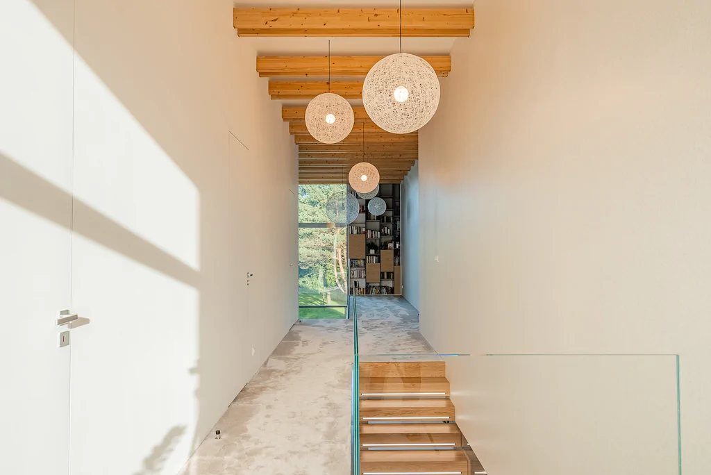 Korytarz na piętrze z drewnianymi belkami, kulistymi lampami i schodami ze szklaną balustradą.