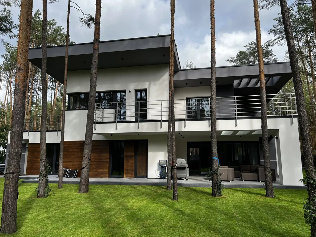 Elewacja ogrodowa: biała fasada z drewnianymi akcentami, balkon i taras z meblami na tle lasu