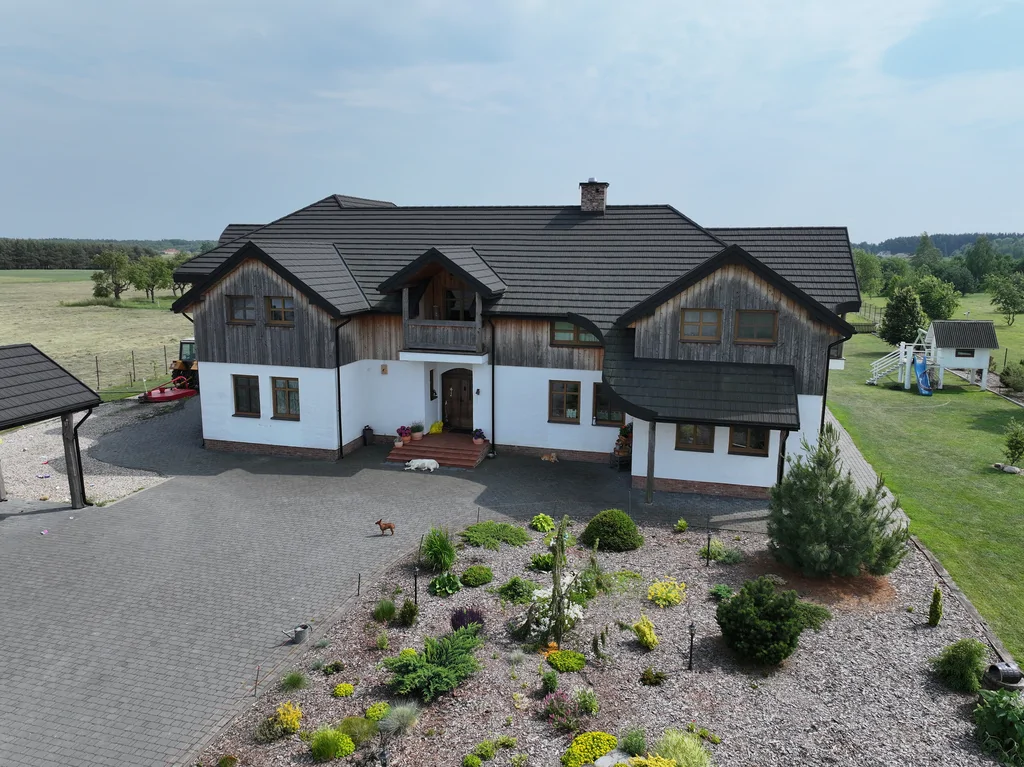 Front domu z drona z rabatą żwirową i roślinami ozdobnymi; na podjeździe widoczne psy