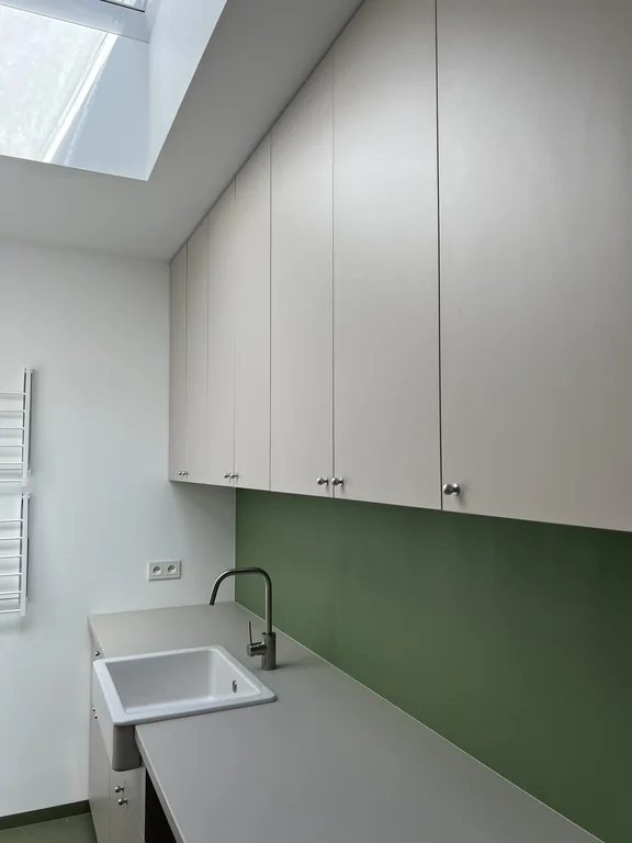 Zabudowa w pomieszczeniu gospodarczym z białymi szafkami, zieloną ścianą i umywalką