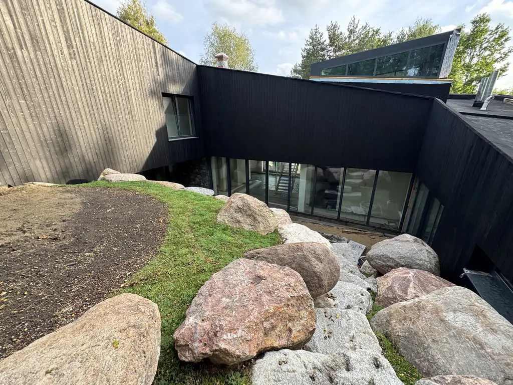 Taras przy czarnej drewnianej elewacji domu z dużymi przeszkleniami i oporową skarpą wykończoną głazami.