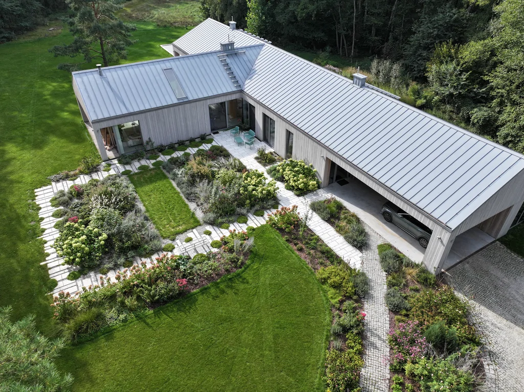 Widok z drona na parterowy dom w kształcie litery L: elewacja drewniana, dach na rąbek i wewnętrzny dziedziniec z ogrodem oraz ścieżkami z płyt.