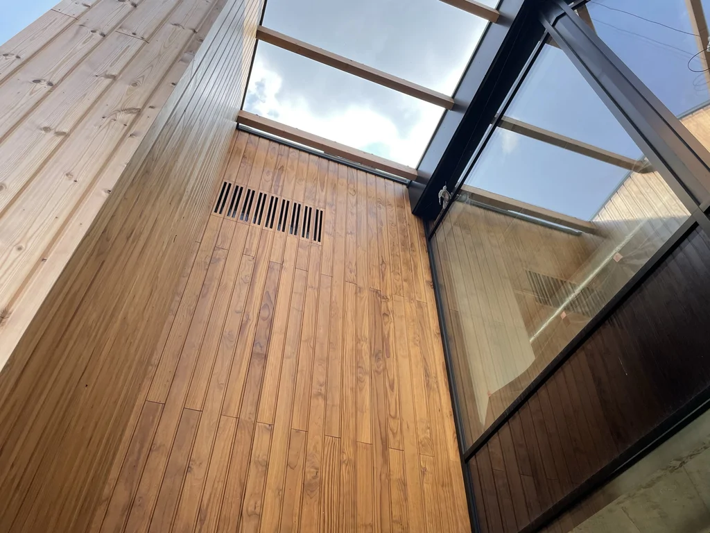 Przeszklone atrium z drewnianą okładziną ścian – widok w górę na świetlik dachowy i połączenie szkła ze stalą.