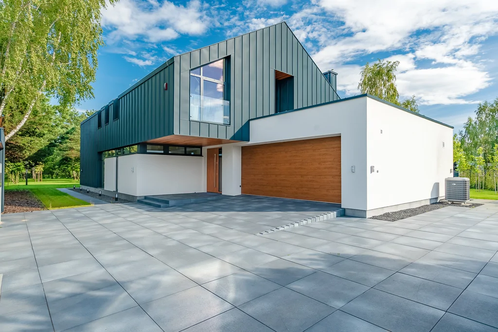 Frontowa elewacja nowoczesnego domu z dużym podjazdem, garażem i nadwieszoną bryłą piętra.