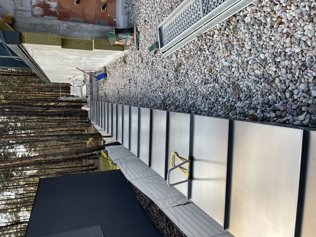 Boczny ciąg komunikacyjny przy domu z metalowym wykończeniem, żwirem i elewacją w trakcie prac