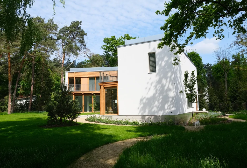 Widok z ogrodu na boczną elewację domu z białą ścianą, zakrzywioną ścieżką i częścią drewnianej zabudowy