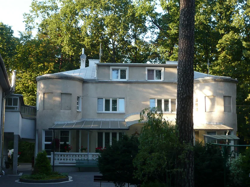 Elewacja frontowa budynku po remoncie latem; zaokrąglone narożniki i drzewa w tle.