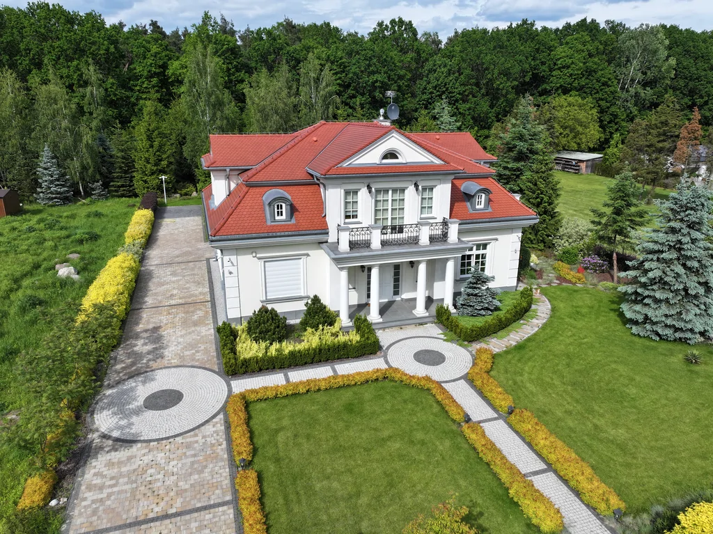 Widok z drona na dom jednorodzinny w stylu dworkowym z białą elewacją z boniami, kolumny okrągłe w strefie wejścia, na balkonie również kolumny i stalowa barierka, pokrycie dachu karpiówka, na dachu widoczne lukarny, przy domu kostka, działka otoczona drzewami 