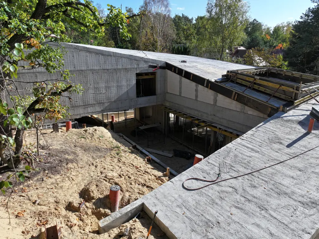 Pionowy widok z drona na budowę nowoczesnego domu z żelbetową konstrukcją, centralnym otworem patio i pochyłym układem dachu.