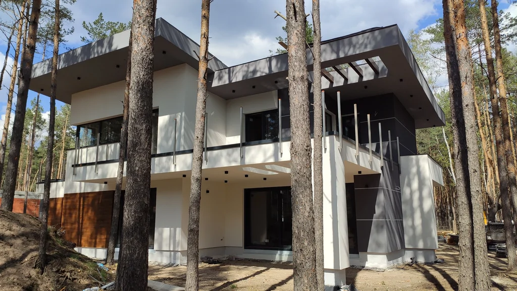 Boczna elewacja gotowego domu z dużymi okapami oraz tarasem/balkonem, biel i grafit wśród sosen