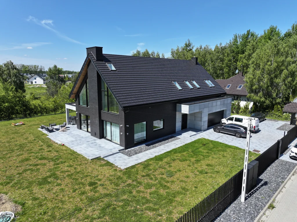 Ujęcie z drona: widok frontowo-boczny domu w Ruścu, dach dwuspadowy ze świetlikami i garaż w jasnej okładzinie.