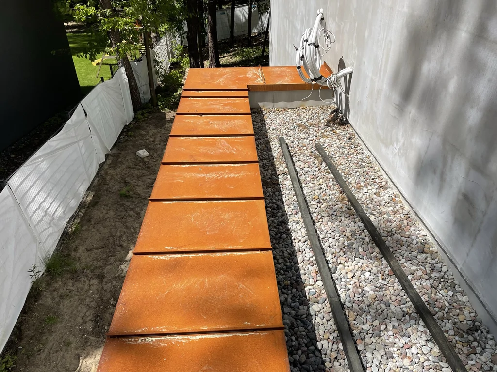 Boczne dojście przy domu z cortenowymi płytami, żwirową opaską i elewacją w trakcie wykańczania