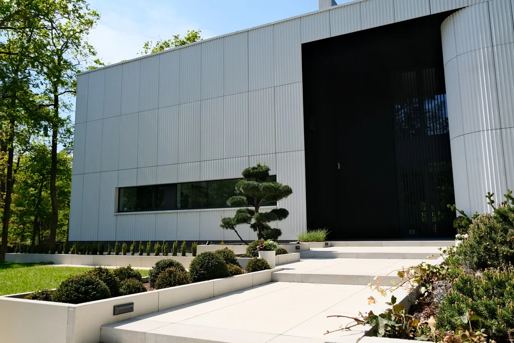 Wejście do nowoczesnego domu z szerokimi schodami, minimalistycznym ogrodem i jasną elewacją ryflowaną