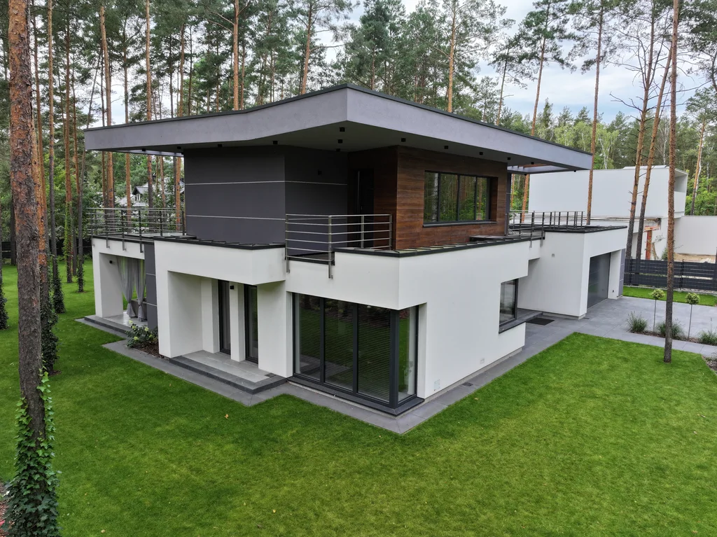 Ujęcie z drona od strony ogrodu: płaski dach i tarasy, bryła w bieli, graficie i drewnie wśród sosen