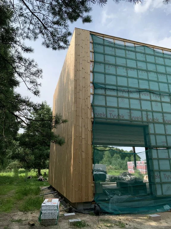 Elewacja boczna w drewnie; pozostała część fasady zabezpieczona membraną i siatką, przygotowanie pod duże przeszklenie.