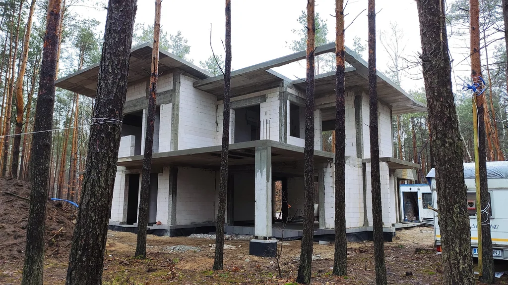 Dwukondygnacyjny dom w stanie surowym z płytami stropowymi, widok przez drzewa na działce leśnej