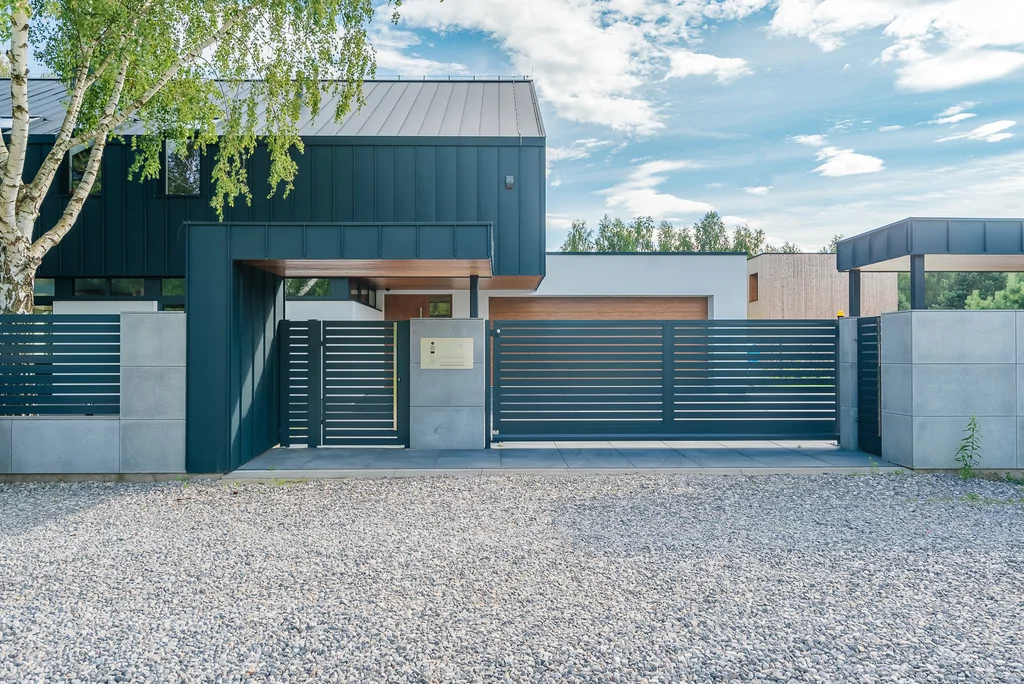 Front posesji nowoczesnego domu z metalowym ogrodzeniem, bramą przesuwną i widoczną elewacją od strony ulicy.