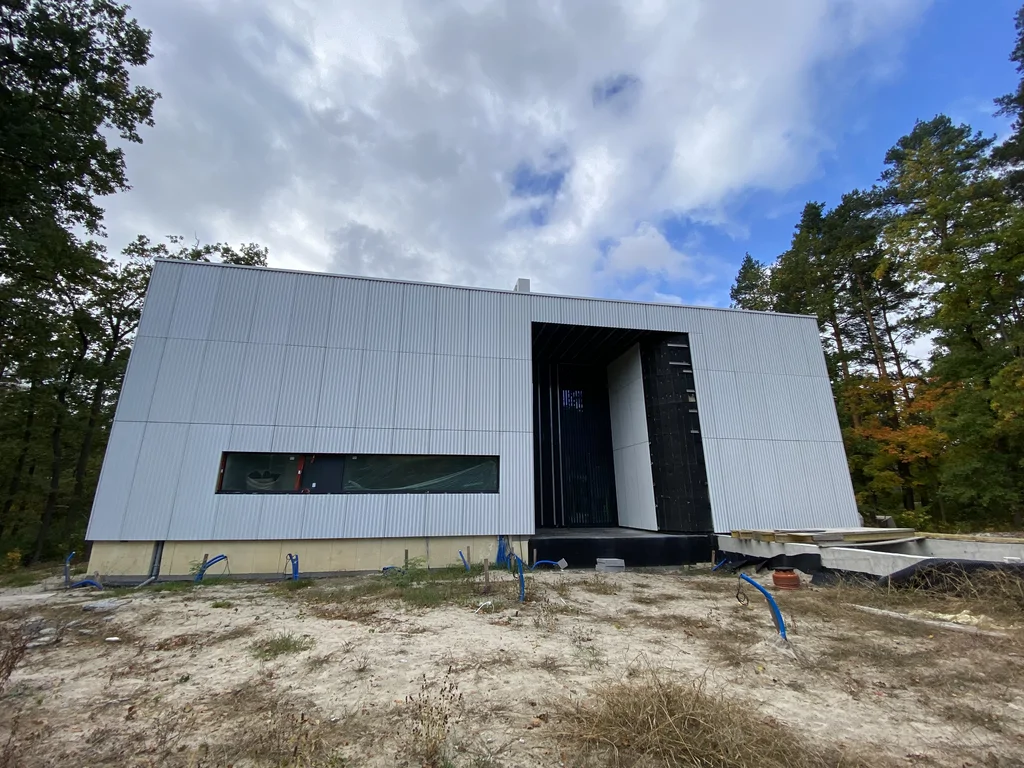 Front nowoczesnego domu w trakcie realizacji z jasną ryflowaną elewacją, cofniętym wejściem i wąskim poziomym oknem