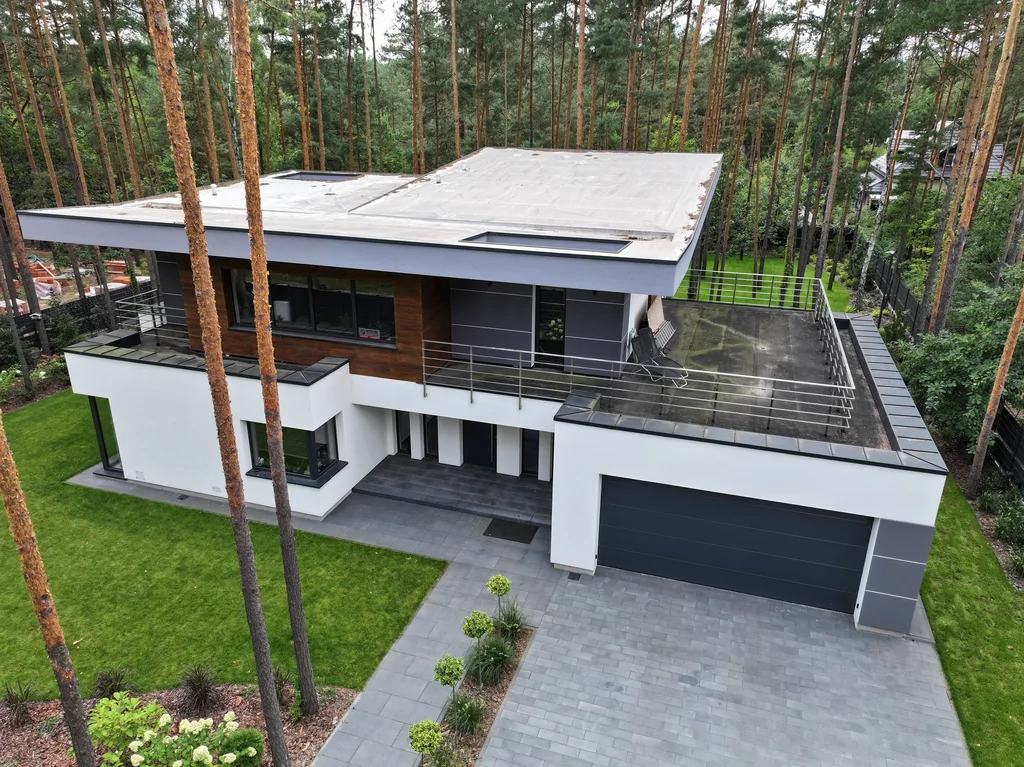 Ujęcie z drona od frontu: nowoczesny dom z płaskim dachem, garażem i dużym tarasem na piętrze wśród sosen