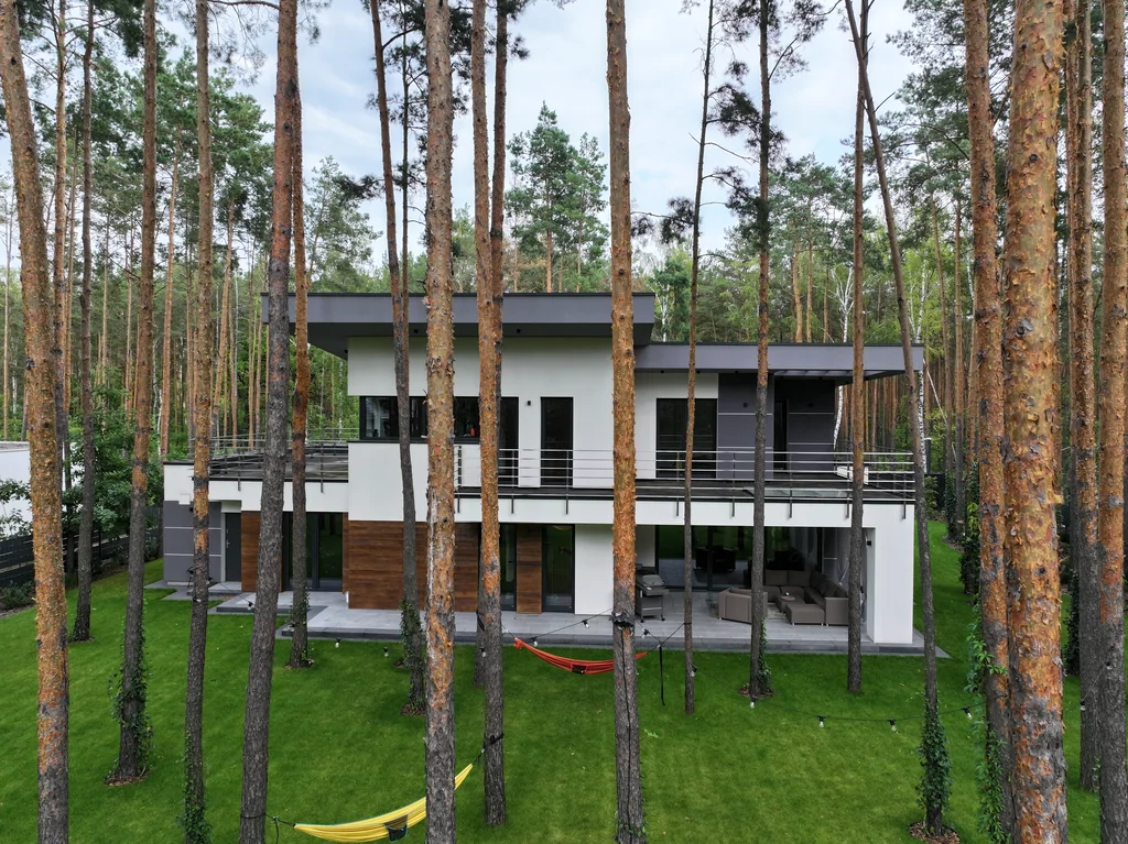 Elewacja ogrodowa z drona: taras pod zadaszeniem i balkon na całej długości, hamaki na trawniku między sosnami