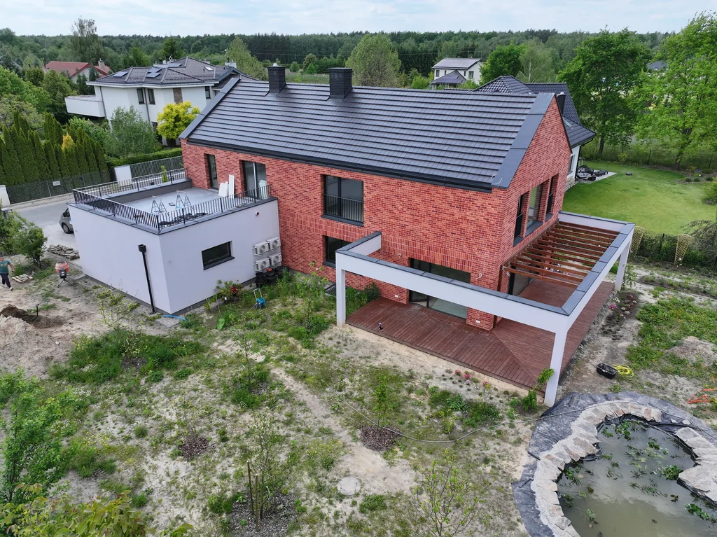 Zbliżenie z drona na tylną część domu z drewnianym tarasem, pergolą i boczną bryłą z tarasem dachowym