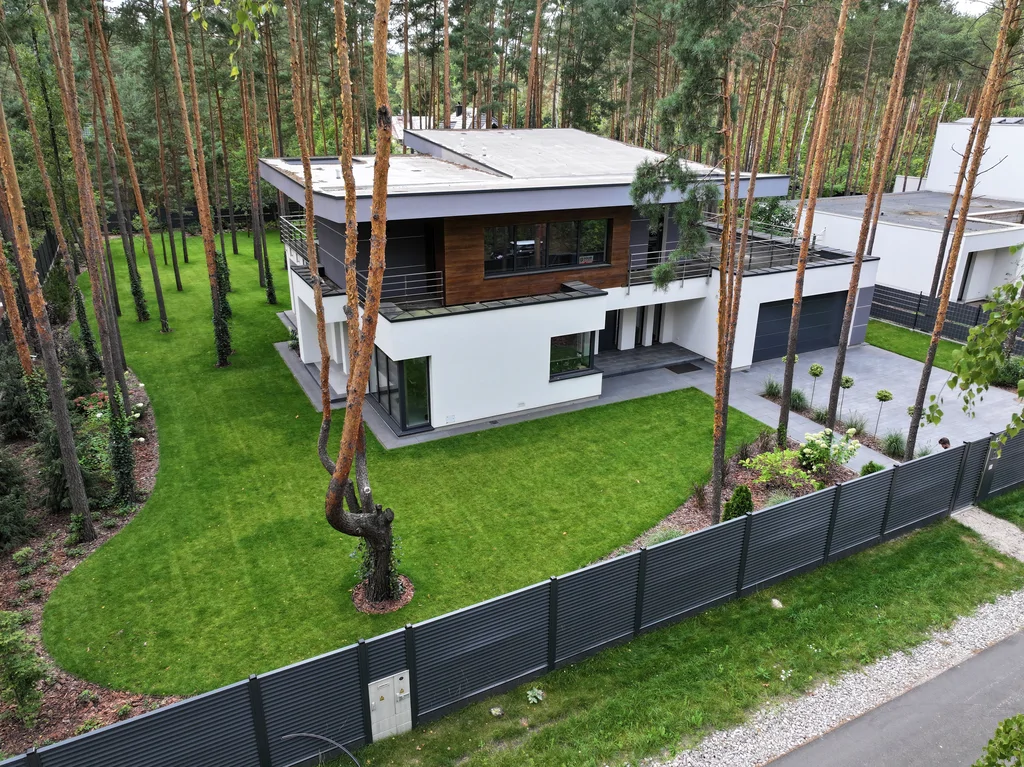 Widok z drona na działkę leśną: dom z dużym trawnikiem i ogrodzeniem lamelowym, bryła w bieli i graficie