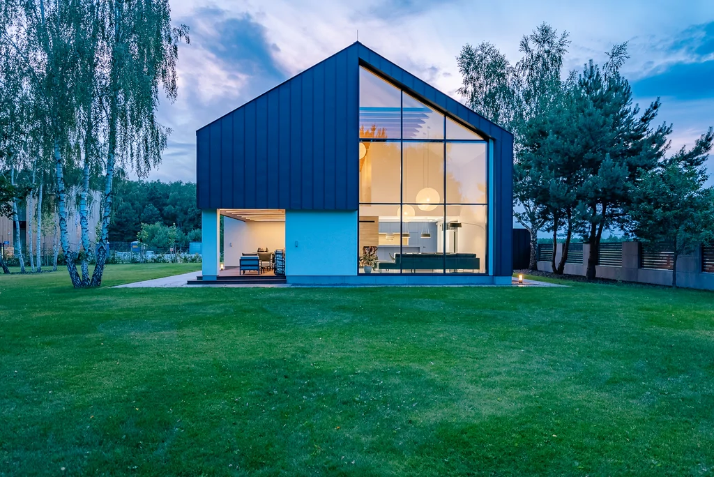 Szczytowa elewacja domu o zmierzchu z dużym przeszkleniem i widocznym, ciepło oświetlonym wnętrzem.