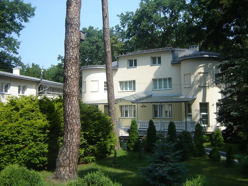 Widok ogrodowy na budynek po remoncie; zaokrąglone skrzydła i przeszklone zadaszenie tarasu.