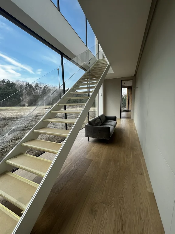 Klatka schodowa z boku: lekkie schody ażurowe, szklana balustrada i widok na zieleń przez duże okno.