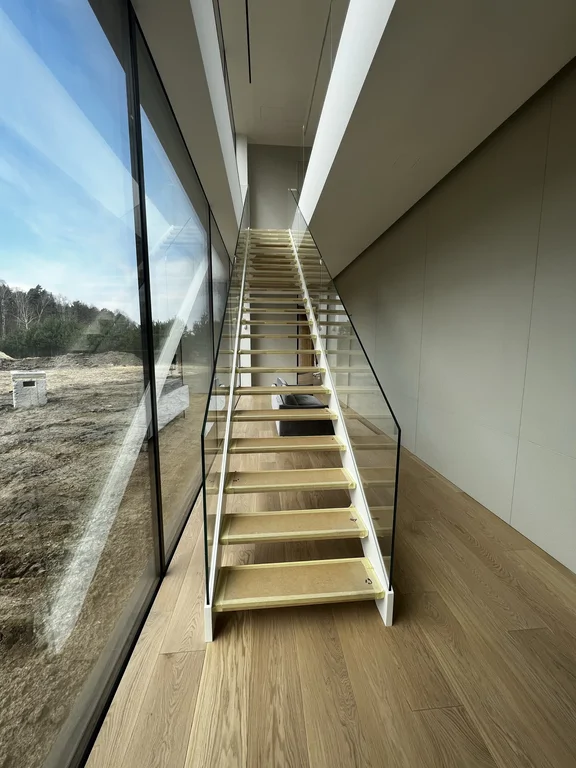 Schody ażurowe ze szklaną balustradą przy dwukondygnacyjnym przeszkleniu i dębowej podłodze.