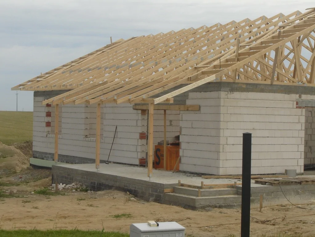 Widok narożnika domu z montowaną więźbą dachową i otworami okiennymi.