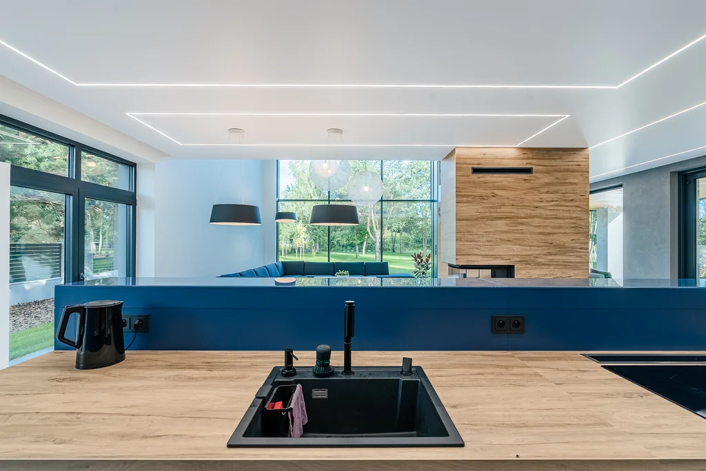 Widok z wyspy kuchennej na salon z kominkiem i wysokimi przeszkleniami.