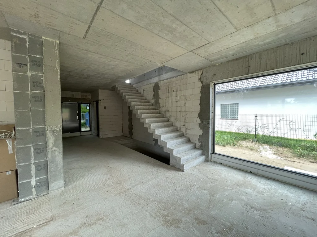 Wnętrze domu w stanie surowym z żelbetowymi schodami, dużym przeszkleniem i otwartą przestrzenią parteru