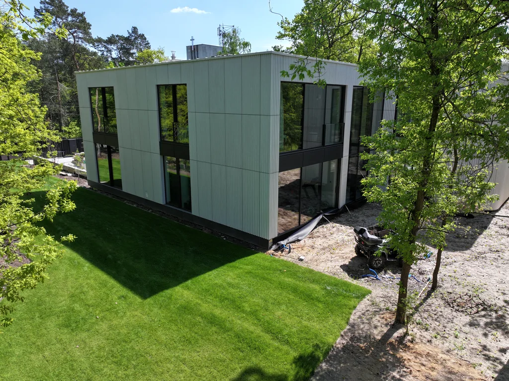 Boczna elewacja nowoczesnego domu z dużymi przeszkleniami, zielonym trawnikiem i drzewami wokół posesji