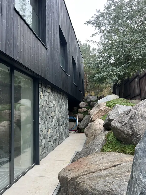 Widok z góry na dziedziniec nowoczesnego domu z czarną elewacją, przeszklonymi ścianami i zieloną skarpą z głazami.