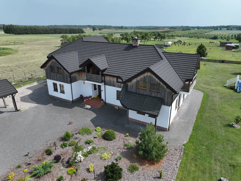 Widok z drona na front domu: wejście główne, zadaszenie i brukowany podjazd oraz rabata z roślinami