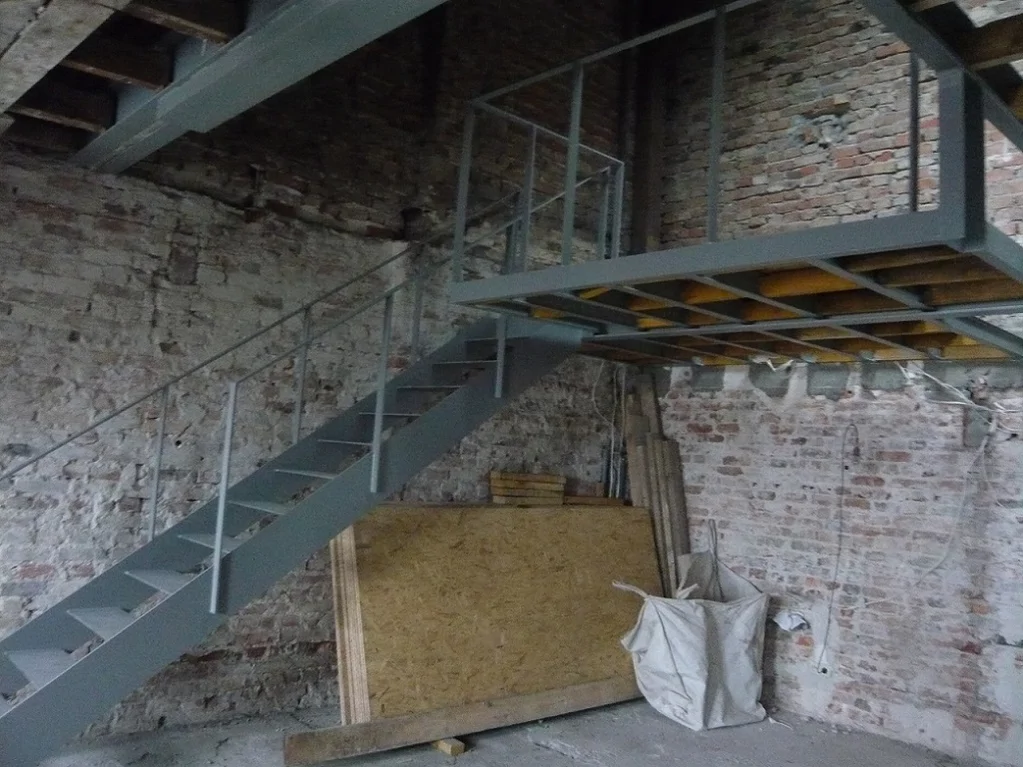 Stalowe schody z poręczą prowadzące na antresolę; surowe ceglane ściany i konstrukcja pod podest