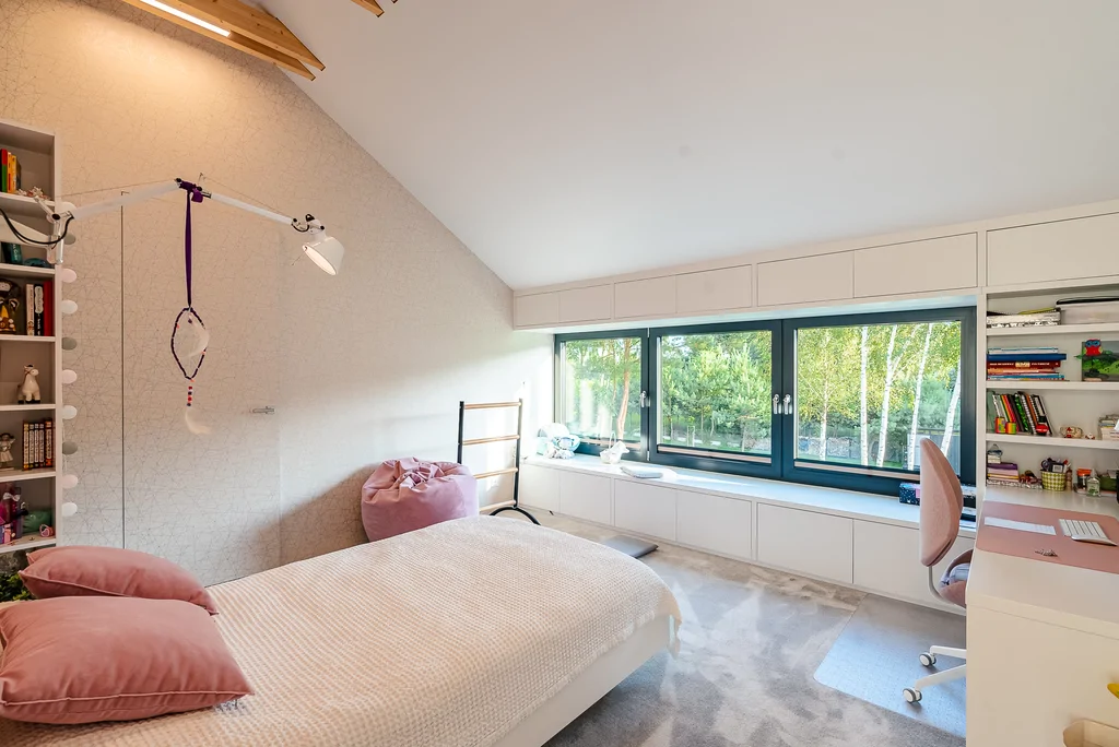 Pokój dziecięcy z różowym łóżkiem, białą zabudową pod oknem i biurkiem przy regale.