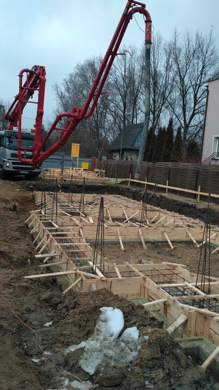 Wylewanie ław fundamentowych, widoczne zbrojenie przed zalaniem i szalunki, w tle pompa do betonu 