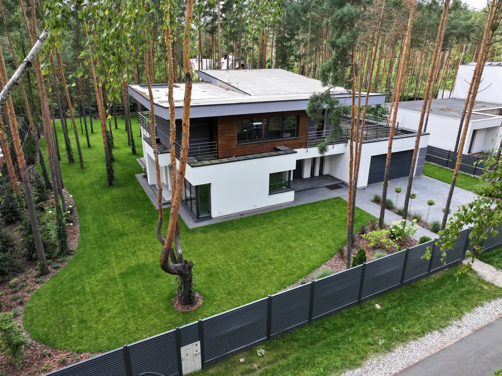 Panorama z drona: dom na ogrodzonej działce w sosnowym lesie, tarasy i płaski dach, widok od ulicy