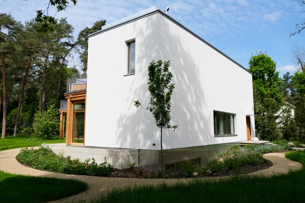 Boczne ujęcie białej bryły domu z drewnianym narożnikiem, ścieżką ogrodową i zielenią wokół budynku