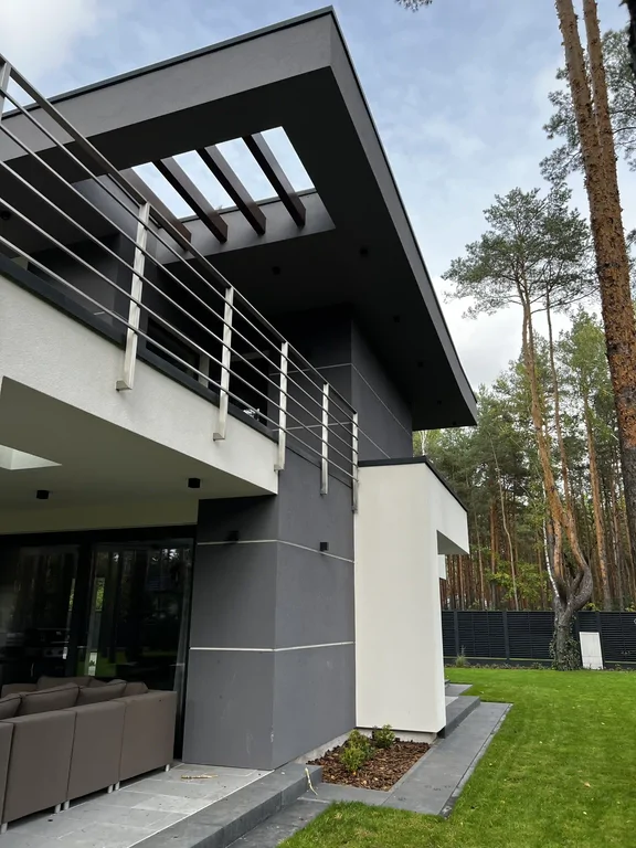 Narożnik budynku z grafitowym pasem i pergolą w okapie; taras i trawnik przy ogrodzeniu