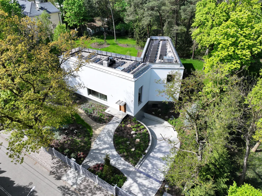 Widok z lotu ptaka na nowoczesny biały dom otoczony drzewami, z zakrzywionym podjazdem wejściowym i panelami fotowoltaicznymi na dachu