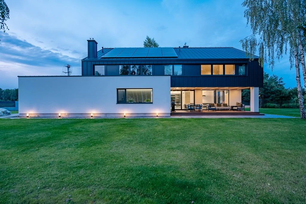 Tylna elewacja nowoczesnego domu o zmierzchu z oświetlonym tarasem i dużym ogrodem.