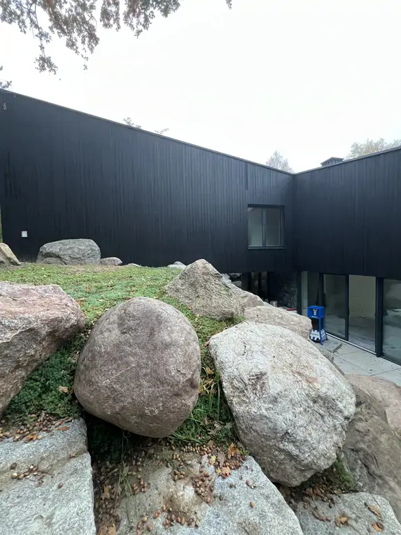 Wąskie przejście wzdłuż elewacji domu z czarnym drewnem, kamiennym wykończeniem i dużymi głazami przy skarpie.